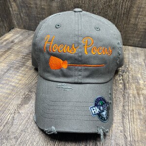 May include: A gray distressed baseball cap with the text "Hocus Pocus" embroidered in orange with a broom and handle. The cap has a black and white BK Caps logo on the side.