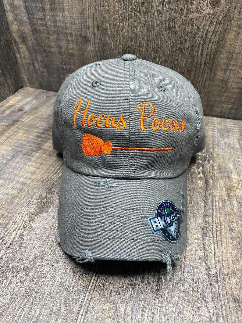 May include: A gray distressed baseball cap with the text "Hocus Pocus" embroidered in orange with a broom and handle. The cap has a black and white BK Caps logo on the side.