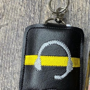 May include: A black faux leather keychain holder featuring a horizontal yellow embroidered stripe and a silver embroidered headset icon. The rectangular holder has rounded corners, black stitching along the edges, and a silver metal lobster clasp.