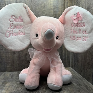 May include: A pink plush elephant with embroidered details. The elephant has large ears with embroidered text and a butterfly. The text reads "Stella Louise Hawkins" and birth details: 7 lbs. 11 oz., 20 inches, and the date and time.