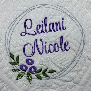 May include: White quilted fabric featuring the name "Leilani Nicole" embroidered in purple script within a grey circular frame. A green and purple floral design is at the bottom of the frame. The fabric has a repeating pattern.