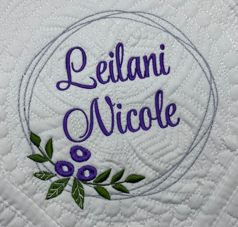 May include: White quilted fabric featuring the name "Leilani Nicole" embroidered in purple script within a grey circular frame. A green and purple floral design is at the bottom of the frame. The fabric has a repeating pattern.