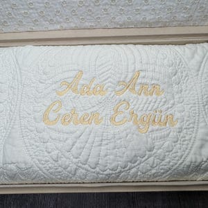 May include: A white, quilted blanket with embroidered gold lettering inside a beige box. The blanket features a detailed, symmetrical pattern and the names "Ada Ann Ceren Ergün" in a cursive font. The box has a patterned interior.