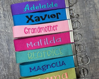 May include: A collection of colorful fabric keychains with embroidered names: "Adelaide", "Xavier", "Grandmother", "Matilda", "Beatrice", "Magnolia", and "Mama". Each keychain features a silver-colored metal ring and a matching fabric loop.