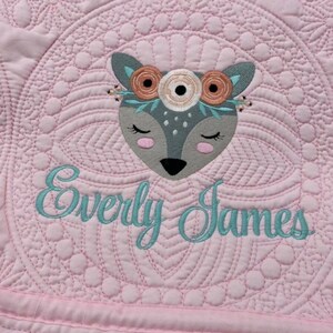 K&ouml;nnte beinhalten: Eine rosafarbene gesteppte Babydecke mit einem gestickten grauen Reh, das eine Blumenkrone tr&auml;gt, und dem Namen "Everly James", der darunter gestickt ist.