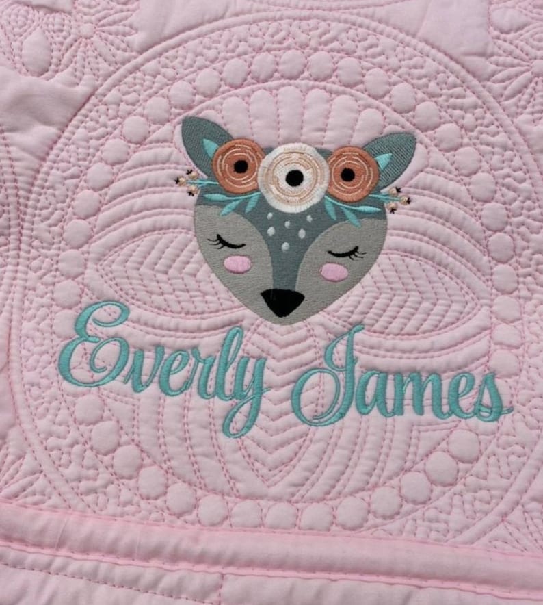 K&ouml;nnte beinhalten: Eine rosafarbene gesteppte Babydecke mit einem gestickten grauen Reh, das eine Blumenkrone tr&auml;gt, und dem Namen "Everly James", der darunter gestickt ist.