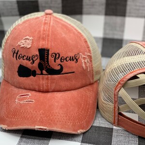 May include: A distressed coral-colored baseball cap with a black mesh back. The front of the cap features a black embroidered design of a witch's boot, broom, and the text "Hocus Pocus".