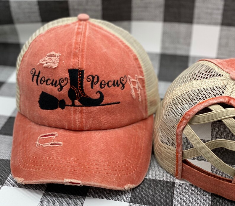 May include: A distressed coral-colored baseball cap with a black mesh back. The front of the cap features a black embroidered design of a witch's boot, broom, and the text "Hocus Pocus".