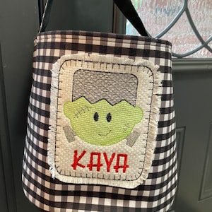 May include: Black and white checkered Halloween trick-or-treat bag with a green Frankenstein face and the name "KAYA" embroidered in red.