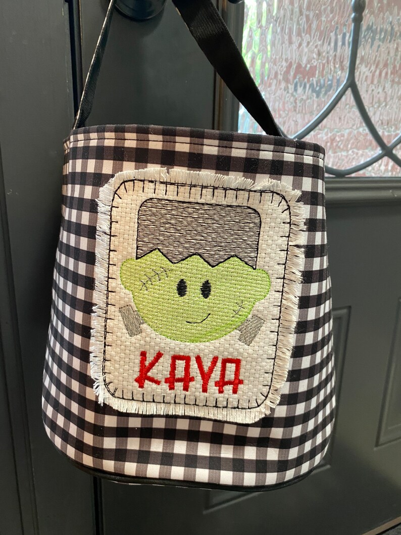 May include: Black and white checkered Halloween trick-or-treat bag with a green Frankenstein face and the name "KAYA" embroidered in red.