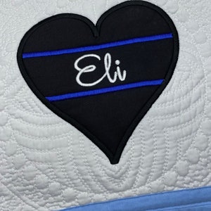 May include: A black heart-shaped patch with a blue line design and the name "Eli" embroidered in white.