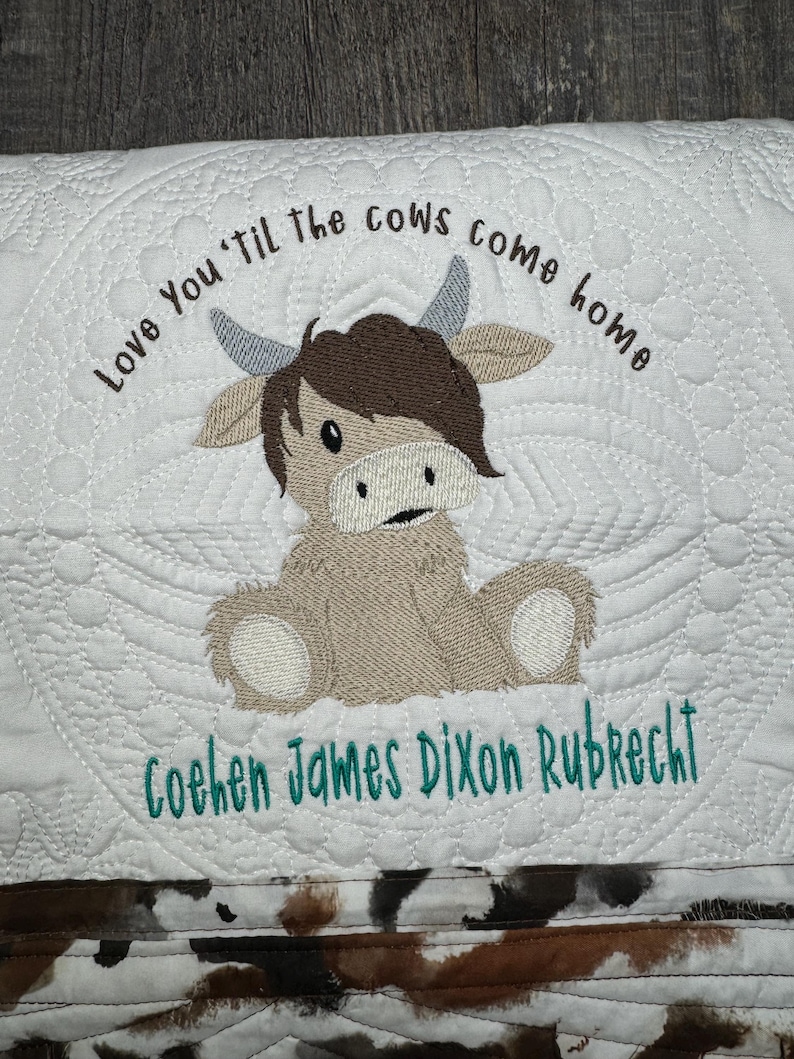 May include: A white quilted baby blanket with a brown cow applique and the text "Love You 'til the cows come home" and "Cohen James Dixon Rubrecht" embroidered in green thread.
