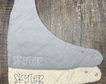 May include: Two baby bibs, one light blue and one cream, are stacked on a wooden surface. Each bib has a bandana shape with snap closures and the name "Skyler" embroidered on it. The bibs are made of a textured fabric.