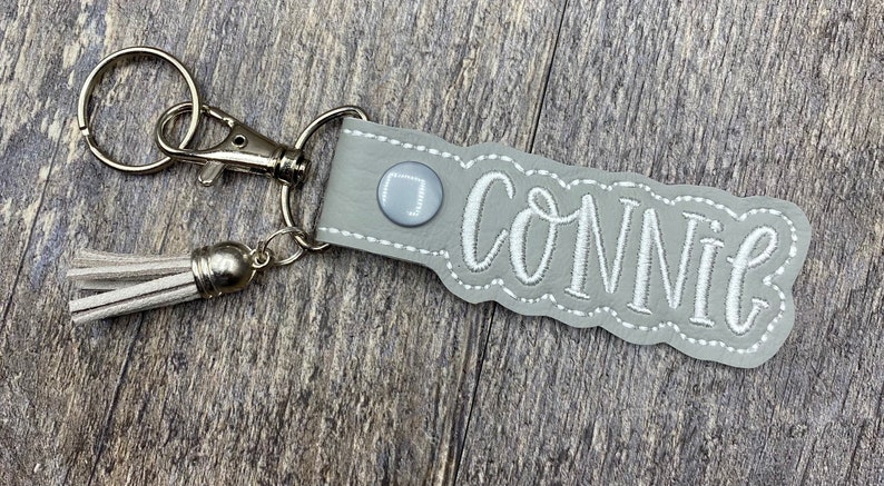 May include: Gray leather keychain with a white tassel and the name "Connie" embroidered in white.