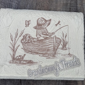 May include: Cream-colored quilted blanket with a brown embroidered design. The design depicts a person fishing from a boat, with a dog, a turtle, and the text "Grandmommy's Threads". The blanket has a decorative stitched border.