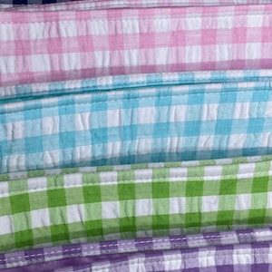 May include: Five fabric swatches in different colors and patterns. The swatches are stacked on top of each other. The top swatch is navy blue with white gingham, the second swatch is pink with white gingham, the third swatch is light blue with white gingham, the fourth swatch is green with white gingham, and the bottom swatch is purple with white gingham.