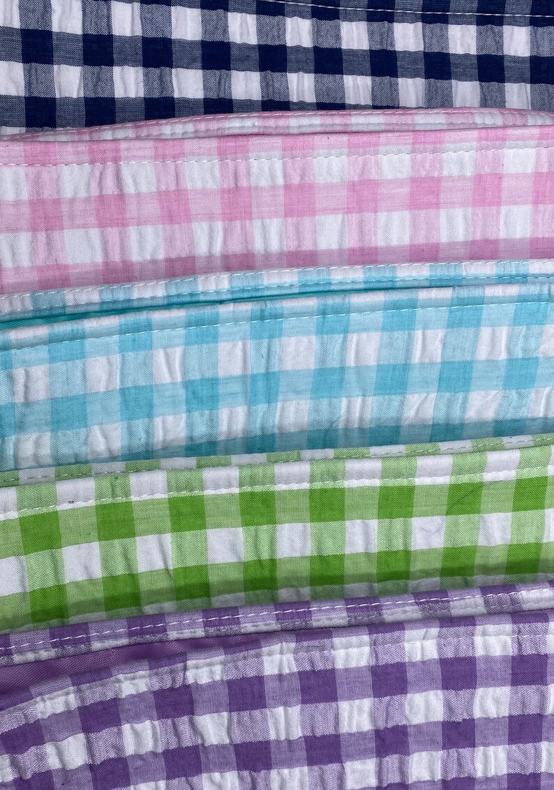 May include: Five fabric swatches in different colors and patterns. The swatches are stacked on top of each other. The top swatch is navy blue with white gingham, the second swatch is pink with white gingham, the third swatch is light blue with white gingham, the fourth swatch is green with white gingham, and the bottom swatch is purple with white gingham.