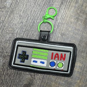 May include: A black and gray video game controller keychain with a green, blue, and red button design. The keychain has a green metal clip and the name "IAN" is embroidered on the controller.