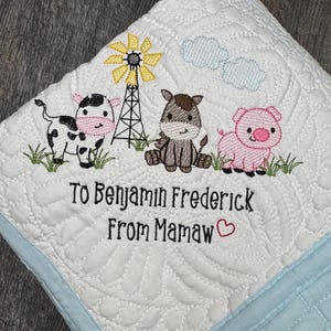 May include: White quilted baby blanket with a light blue border. Embroidered with a farm scene featuring a cow, horse, pig, windmill, and clouds. The text "To Benjamin Frederick From Mamaw" is also embroidered on the blanket.