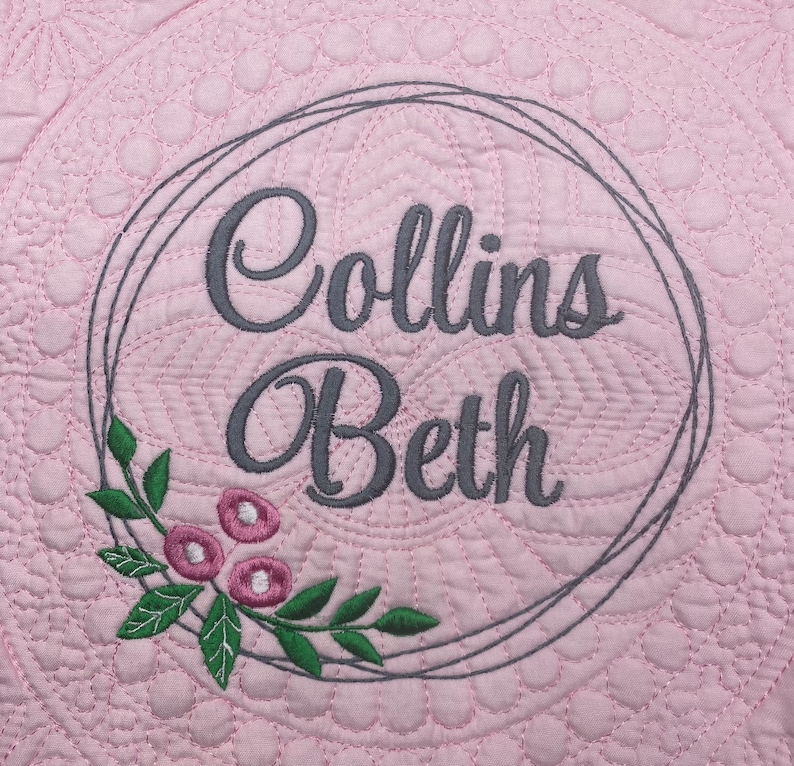 May include: A pale pink quilted fabric with a circular pattern. The centre features the embroidered names "Collins Beth" in grey script within a grey geometric frame, with a floral and leaf design in pink and green.
