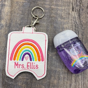May include: White hand sanitizer holder with a rainbow and heart design. The holder has a silver keychain and the text "Mrs. Ellis" is embroidered on the front. A purple hand sanitizer bottle with a unicorn design is in the background.