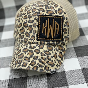 May include: A brown and beige leopard print trucker hat with a black and gold monogram patch that says "KWA".