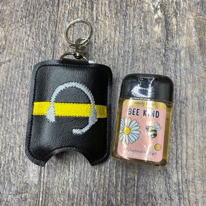 May include: Black leather hand sanitizer holder with a yellow stripe and a white headphone graphic. The holder has a silver keychain attached. A small clear bottle of hand sanitizer with a pink and yellow label that says "simply body BEE KIND" is next to the holder.