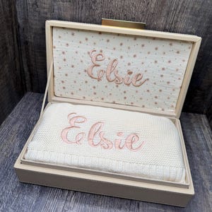 May include: A cream-colored rectangular keepsake box with a hinged lid. The interior lid features a fabric lining with a small floral pattern and the embroidered name Elsie. A folded cream knit blanket with the name Elsie embroidered in pink.