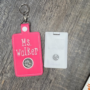 May include: A hot pink key fob with the name "Ms. Walker" embroidered in white. The fob has a silver coin detail and a silver key ring. A white Centegix card is next to the fob.