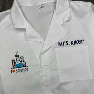 May include: White lab coat with a pocket on the right side. The pocket has the text "Mrs. Kiker" embroidered on it. The left side of the coat has an embroidered design of beakers and the text "I love science".