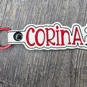 May include: Red and white keychain with a red embroidered name "Corina" and a black silhouette of a running person. The keychain has a red tassel.