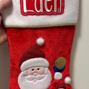 May include: Red and white Christmas stocking with a Santa Claus and reindeer design. The stocking has a white cuff with the name "Eden" embroidered in red.