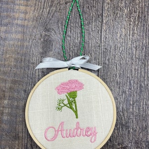 May include: A round wooden embroidery hoop with a white linen fabric background. The hoop features a pink carnation flower with green leaves and the name "Audrey" embroidered in pink thread.