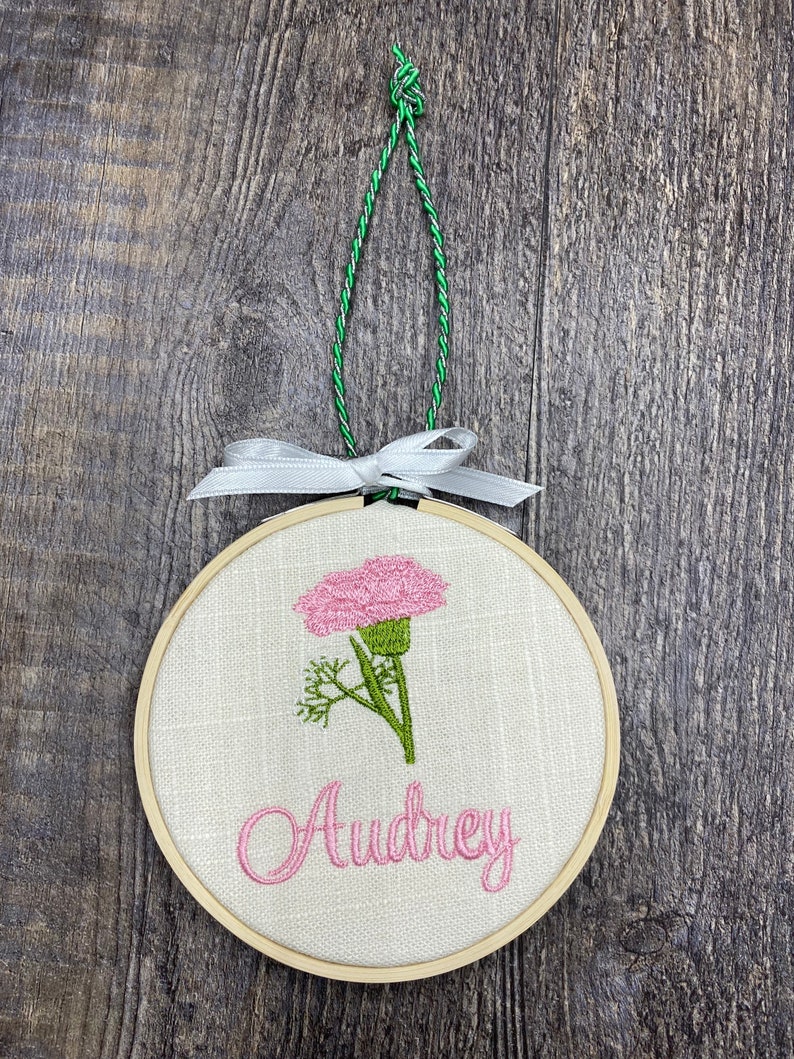 May include: A round wooden embroidery hoop with a white linen fabric background. The hoop features a pink carnation flower with green leaves and the name "Audrey" embroidered in pink thread.