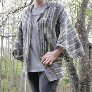 Può includere: Un cardigan a maglia a righe grigie e bianche con cappuccio. Il cardigan ha una vestibilità rilassata e maniche lunghe.