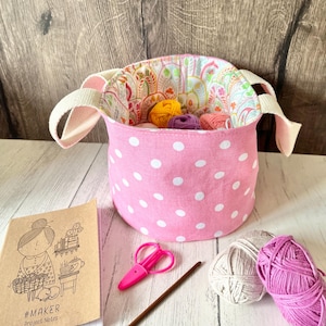 May include: A pink polka-dot fabric basket with white handles, filled with colorful yarn balls. A notebook with a drawing and the text "#MAKER Project Notes" is next to the basket, along with pink scissors and a crochet hook. Yarn craft supplies.