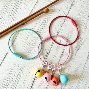 May include: Four colorful jingle bells attached to a silver chain with three silver clasps. The chain is attached to a pink, red, and turquoise cable loop.