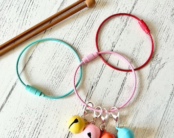 May include: Four colorful jingle bells attached to a silver chain with three silver clasps. The chain is attached to a pink, red, and turquoise cable loop.