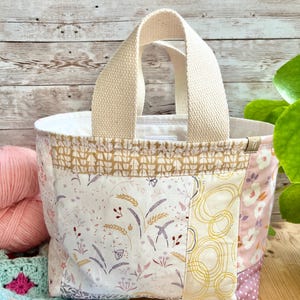 May include: A small fabric tote bag featuring a patchwork design with various floral, wheat, and abstract circular patterns in shades of cream, pink, and mustard yellow. The bag has two thick, cream-colored woven handles and a small side tag.