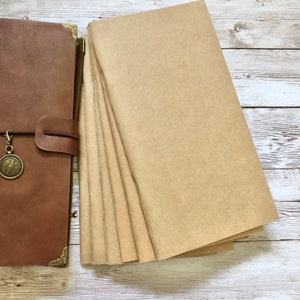 May include: A brown leather journal with a compass charm and a stack of blank kraft paper inserts. The journal has brass corner protectors and a leather strap closure. The paper inserts are stacked next to the journal.