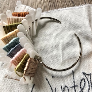 May include: A selection of embroidery threads in a range of colours, attached to a white plastic organiser ring. The threads include white, brown, green, and pink. The organiser is on white fabric with the word "Winter" embroidered in dark grey thread.