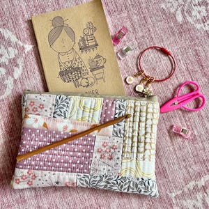 May include: A rectangular quilted pouch featuring a patchwork of floral, polka dot, and leaf patterns in shades of pink, white, and tan. A wooden crochet hook rests on the pouch. Nearby are a tan notebook, a red stitch marker, and pink scissors.
