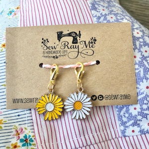 Puede incluir: Dos marcadores de punto de costura en forma de margarita de color dorado con esmalte blanco y amarillo. Los marcadores están unidos a una tarjeta marrón con el texto "Sew Ray Me a handmade life" y la dirección del sitio web "www.sewrayme.com".