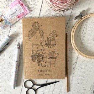 May include: A brown notebook with a cute illustration of a person holding yarn and a cat. The notebook has the text "#MAKER Project Notes" printed on the cover.