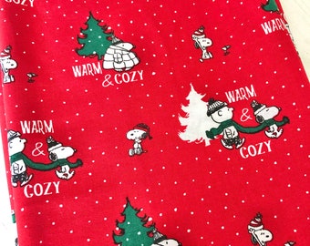 May include: A red fabric featuring a repeating pattern of Snoopy and Charlie Brown characters in winter attire, green Christmas trees, and white snowflakes. The text WARM & COZY is printed in white throughout the design, alongside small illustrations of the characters wearing hats and scarves.