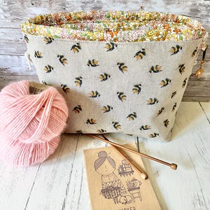 Könnte beinhalten: Eine beigefarbene Stofftasche für Handarbeitsprojekte mit einem wiederkehrenden Hummelmuster und floralem Innenfutter, dazu ein Knäuel rosa Garn, zwei Stricknadeln aus Holz und ein beiges Notizbuch mit dem Text #MAKER Project Notes auf dem Einband.
