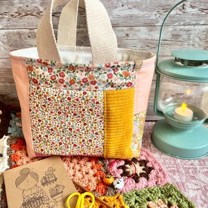 May include: A small fabric tote bag featuring a patchwork design of floral patterns and solid mustard yellow corduroy, with two beige woven handles. The bag is constructed with light pink side panels and a white interior lining.