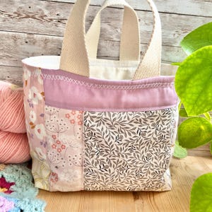 May include: A small fabric tote bag featuring a patchwork design with panels of floral patterns in shades of pink, white, and black. The bag has two cream-coloured canvas handles and a lavender fabric band across the top with white cross-stitch detailing.