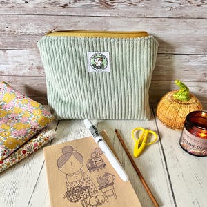 May include: A pale green corduroy pouch with a yellow zip, a small patch, and a notebook with a drawing of a woman. Also, a crochet pumpkin, scissors, a pen, and a candle.