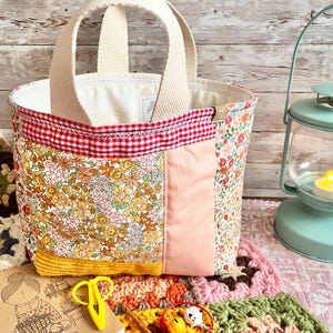 May include: A patchwork tote bag featuring floral fabric panels, a solid pink section, and a yellow corduroy base. The bag has cream-coloured fabric handles and a red and white gingham trim along the top edge, with a small tag on the side.